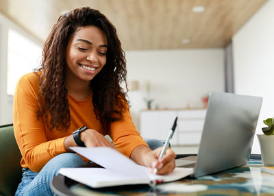 young woman sitting on the couch with a laptop in front of here while she writes on a piece of paper