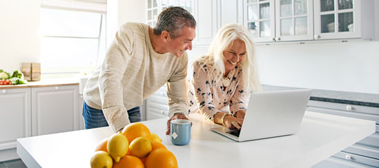 Man and woman standing in their kitchen smiling while looking at their computer
