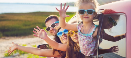 Family waving out of car windows while wearing sunglasses at a beach